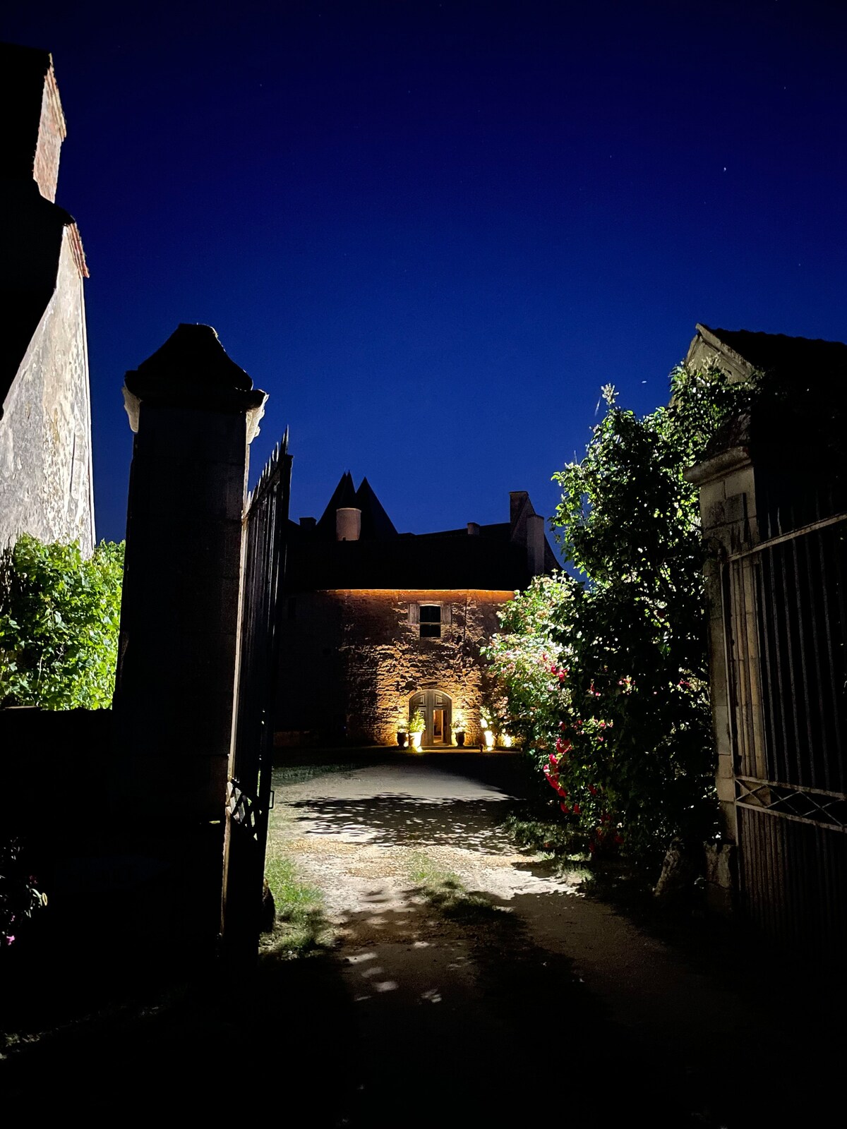 A softly lit pathway leads to the entrance of the historic house, surrounded by lush greenery. The stone façade is subtly illuminated, highlighting architectural details under the night sky, while shadows dance along the ground from the surrounding trees and plants.