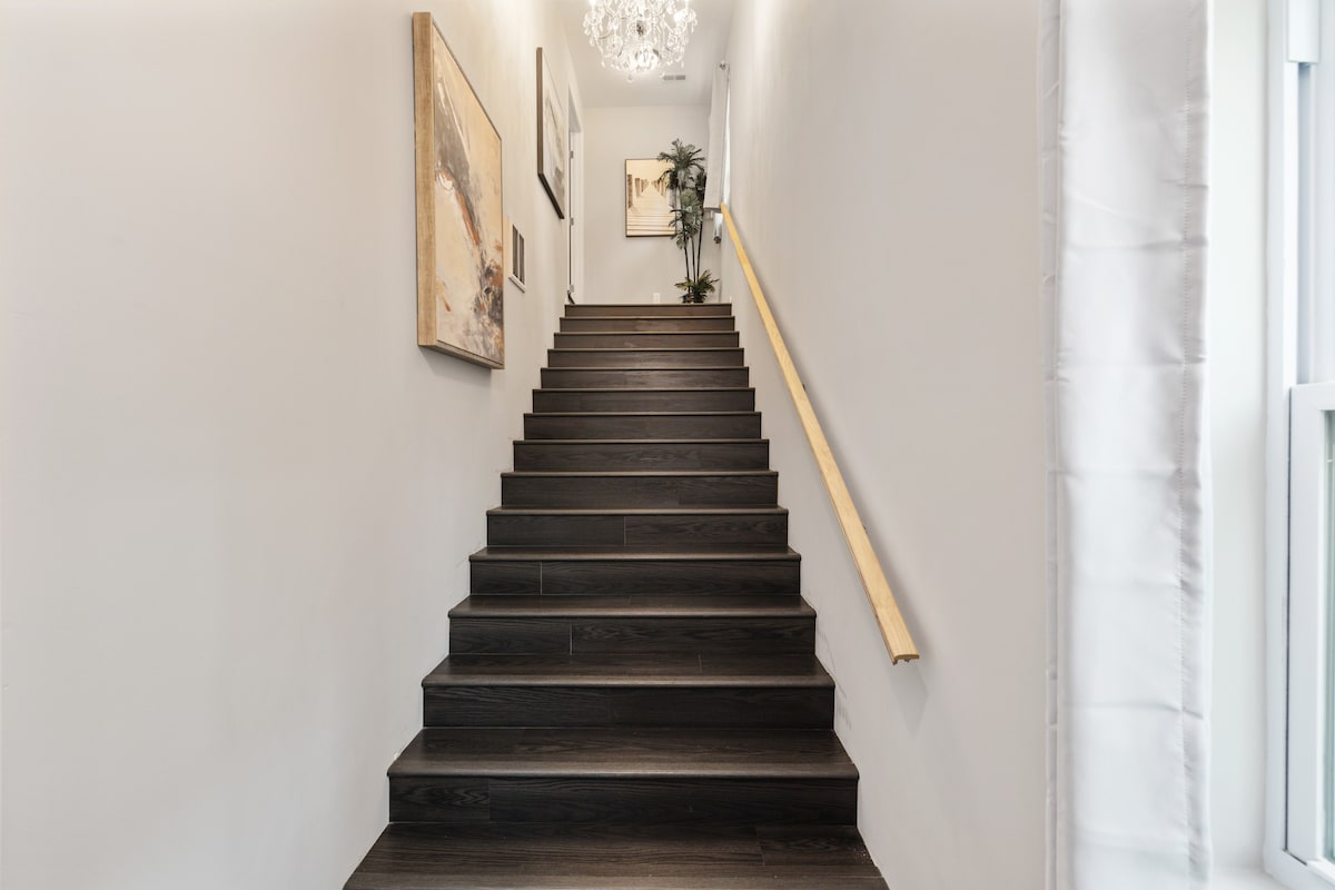 A wide staircase leads to the second floor, featuring dark wood steps and a light-colored handrail. The walls are adorned with artwork, and a modern chandelier hangs from the ceiling. A tall plant is positioned near the top of the stairs.