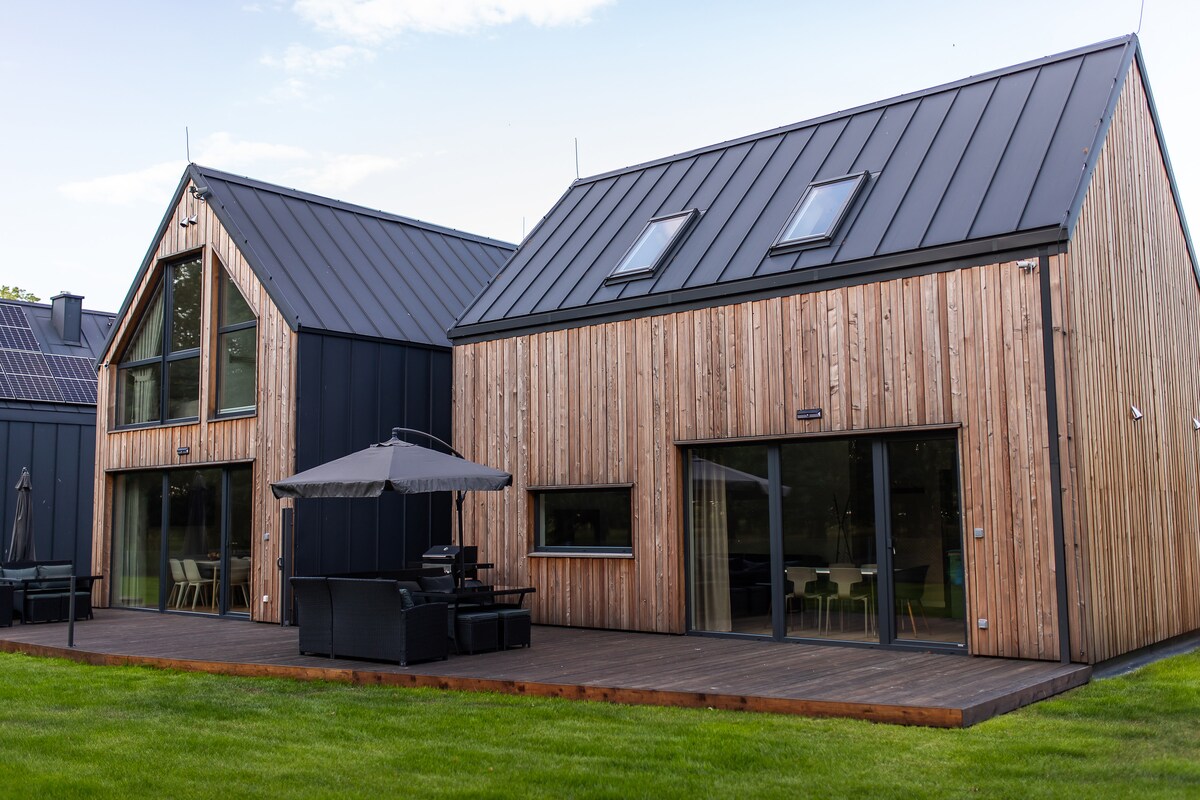 Two modern, two-story cabins are presented with wooden exteriors and metal roofs. Each cabin features large windows that allow natural light to fill the spaces. A spacious deck with outdoor seating is visible, leading to a landscaped grassy area.