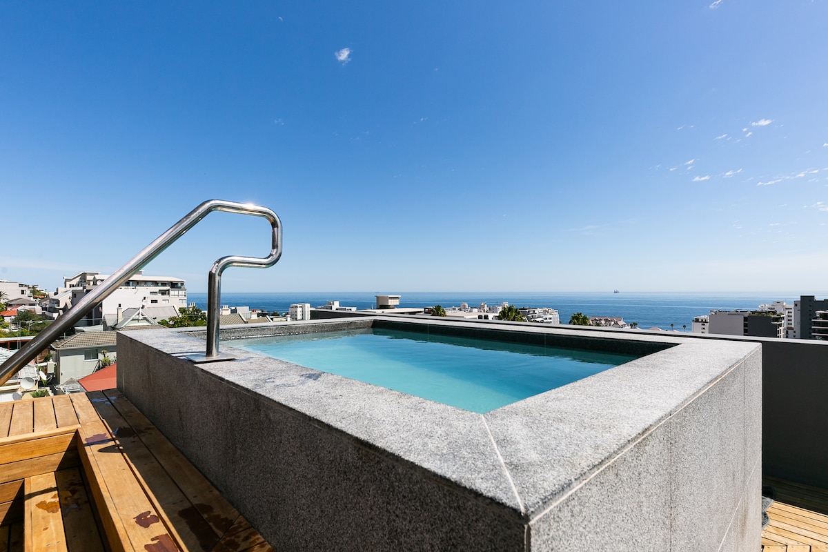 A private jacuzzi is set on a spacious terrace, surrounded by a wooden deck. Clear views of the ocean are visible in the background, complemented by a bright blue sky with a few scattered clouds.