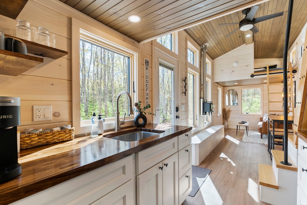 A bright kitchen area features modern white cabinetry and a wooden countertop. Large windows provide natural light and views of the surrounding woods. A small coffee maker is positioned on the counter, with a sink and minimalist decor enhancing the space.