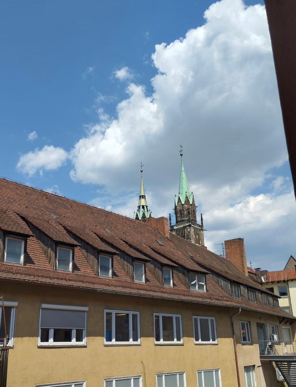 The image captures a view of rooftops in the foreground, featuring a mix of brown and yellow hues. In the background, church towers with green spires rise against a bright blue sky adorned with fluffy white clouds.