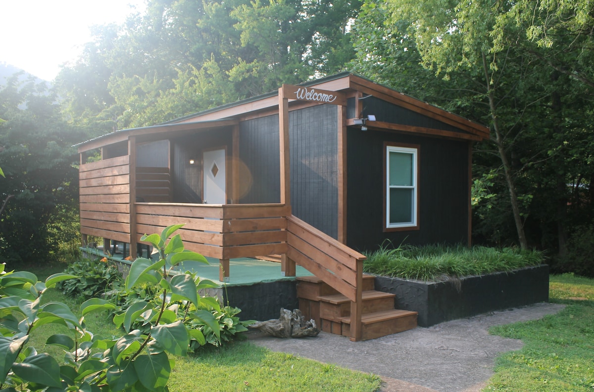 The exterior of the cabin is viewed from the front, featuring a wooden porch with steps leading to the entrance. Green foliage surrounds the structure, complementing the warm wooden tones of the cabin, and a sign with 'Welcome' is visible above the door.
