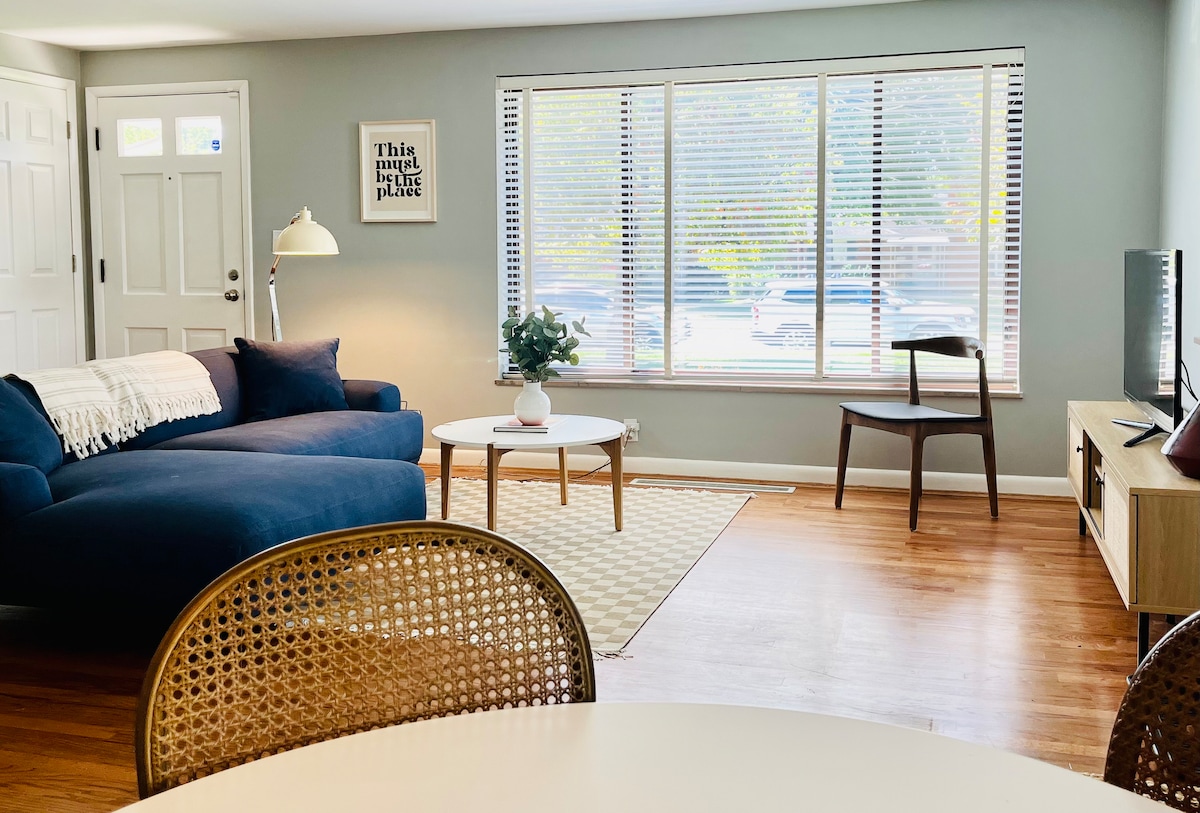 A modern living area features a dark blue sectional sofa paired with a round coffee table. A large window allows natural light to fill the space, while a cozy area rug defines the seating arrangement. A small chair and lamp complete the inviting atmosphere.