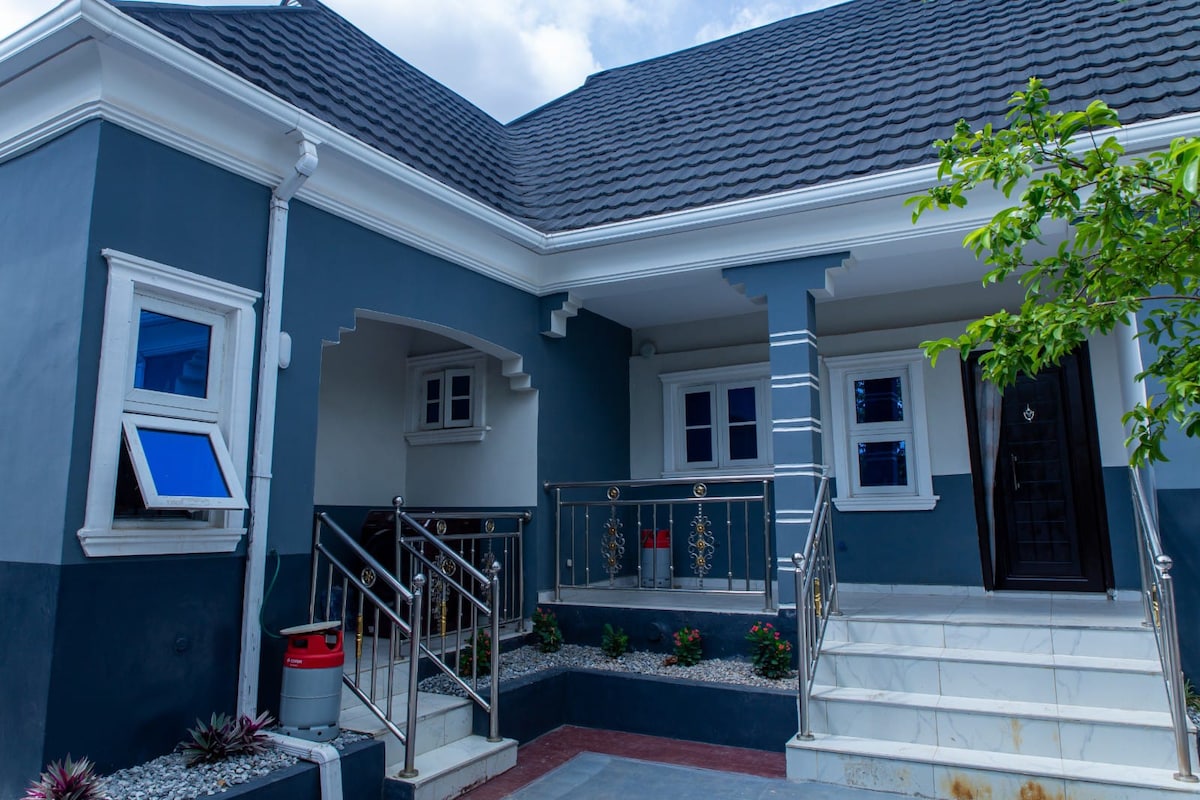 The exterior of the apartment features a modern design with a mix of grey and white accents. A staircase leads to the entrance, framed by decorative railings. Windows are featured on either side, and landscaping includes small stones and potted plants, enhancing the welcoming appearance.