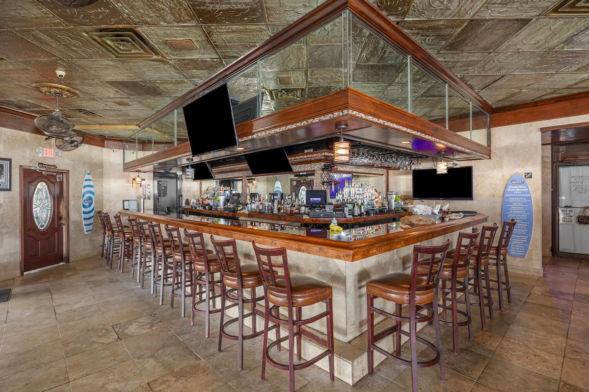 A spacious bar area features a centrally located wooden counter with ample seating around it. The bar is well-stocked with various bottles and glassware, and warm lighting creates a welcoming ambiance. Decorative elements include a textured ceiling and a unique glass partition above the counter.
