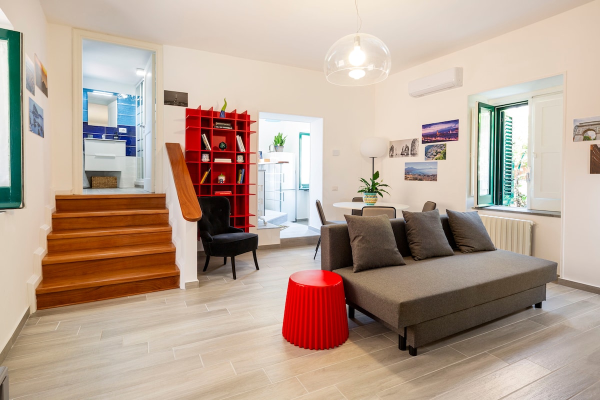A modern living area is presented with a comfortable gray sofa and a bright red side table. The space includes a stylish chair, steps leading to an upper level, and large windows allowing natural light. Decorative elements can be seen on the walls.