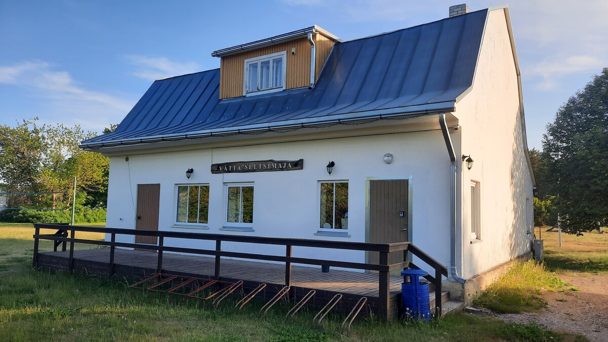 The exterior of a two-story building is displayed, featuring a classic white façade with a dark metal roof. A wooden deck extends from the front, and multiple windows provide natural light. The surrounding grassy area enhances the quiet setting, with trees partially visible in the background.