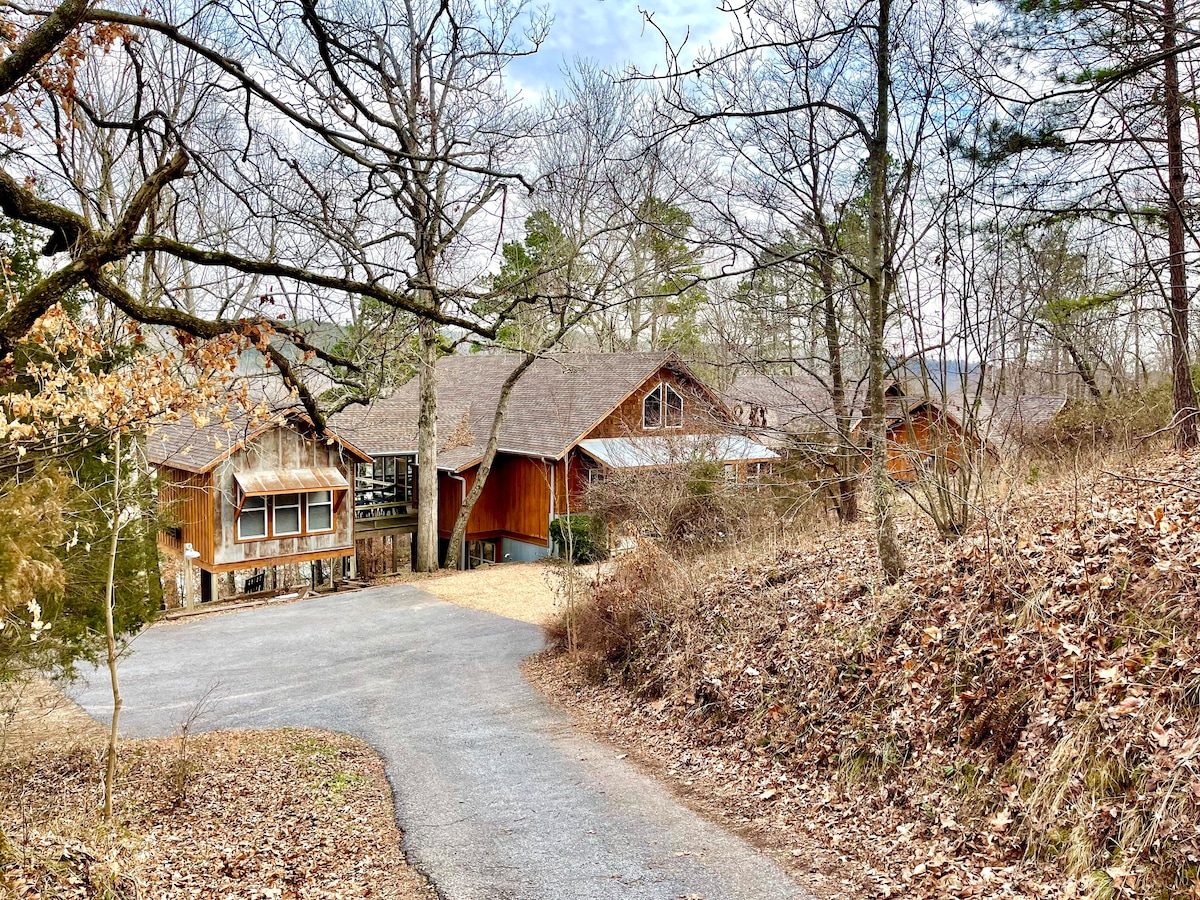 A winding gravel driveway leads to a cluster of wooden structures nestled among leafless trees. The landscape is lightly covered with fallen leaves, showcasing a serene and natural setting, with hints of greenery suggesting the approach of spring.