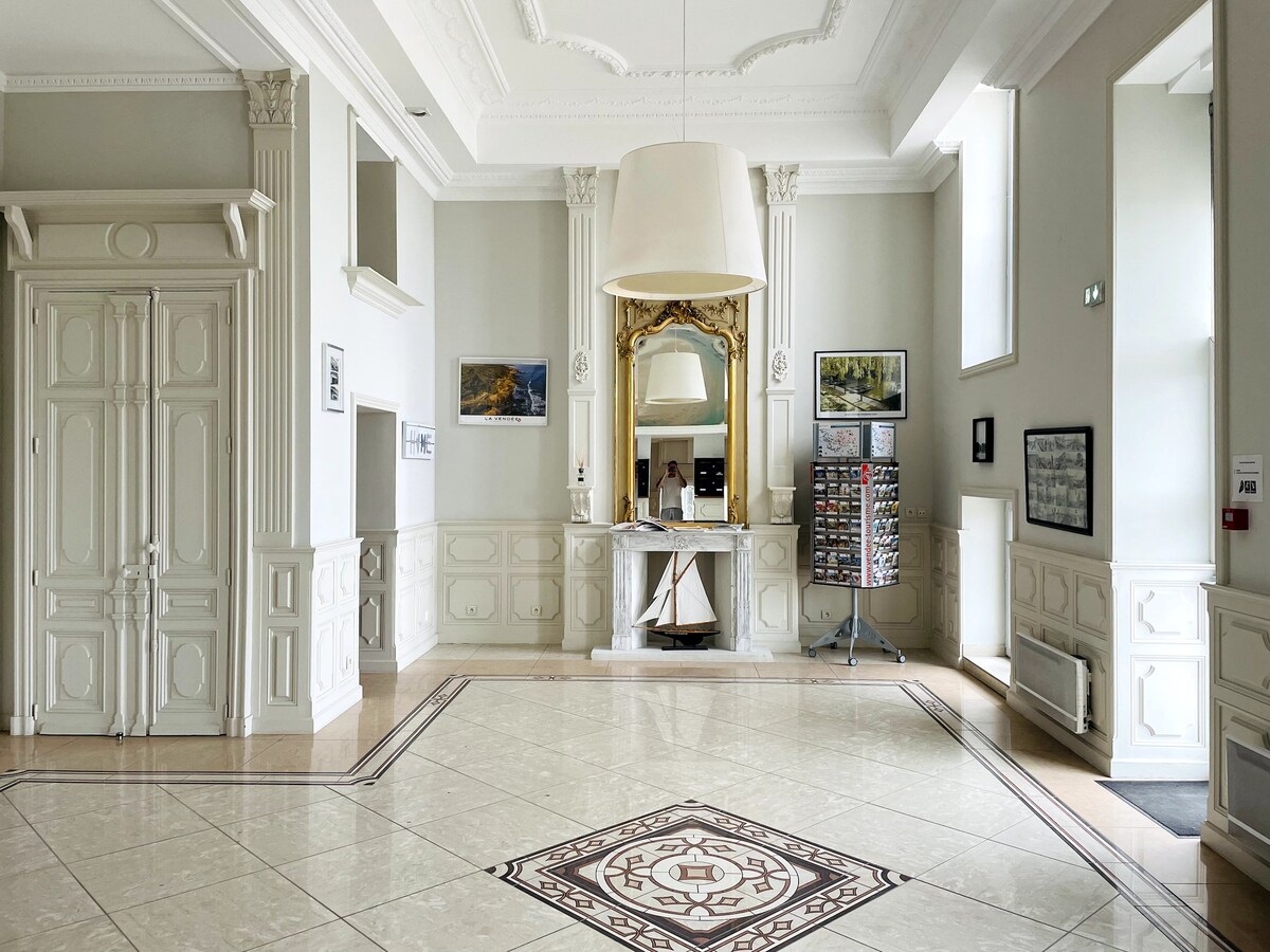 A bright and airy lobby features high ceilings and elegant architectural details, including a large chandelier. The well-lit space includes reception areas, decorative wall art, and a display stand for brochures. A polished tiled floor with an intricate design complements the overall aesthetic.