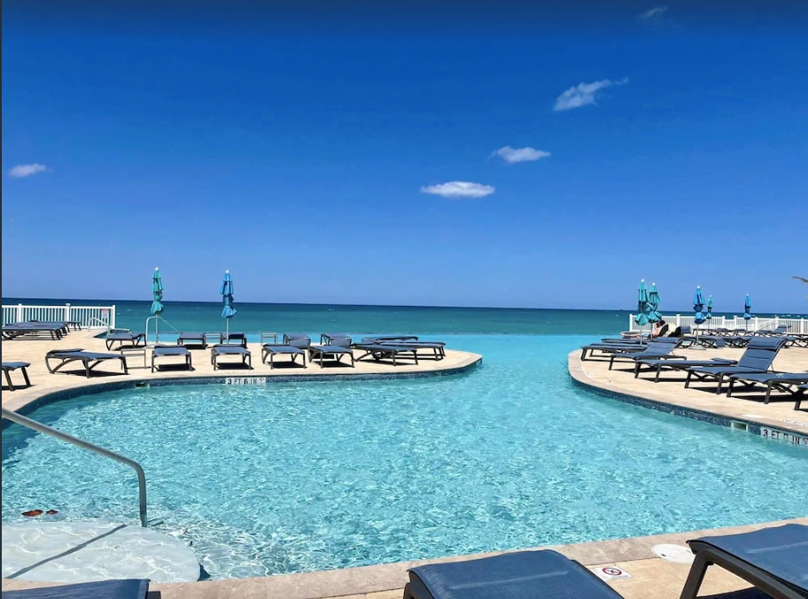 A serene infinity pool merges with the clear blue ocean, creating a seamless horizon. Surrounding the pool, numerous lounge chairs are positioned for guest relaxation. The bright sky reflects off the water, adding to the tranquil atmosphere of the resort.