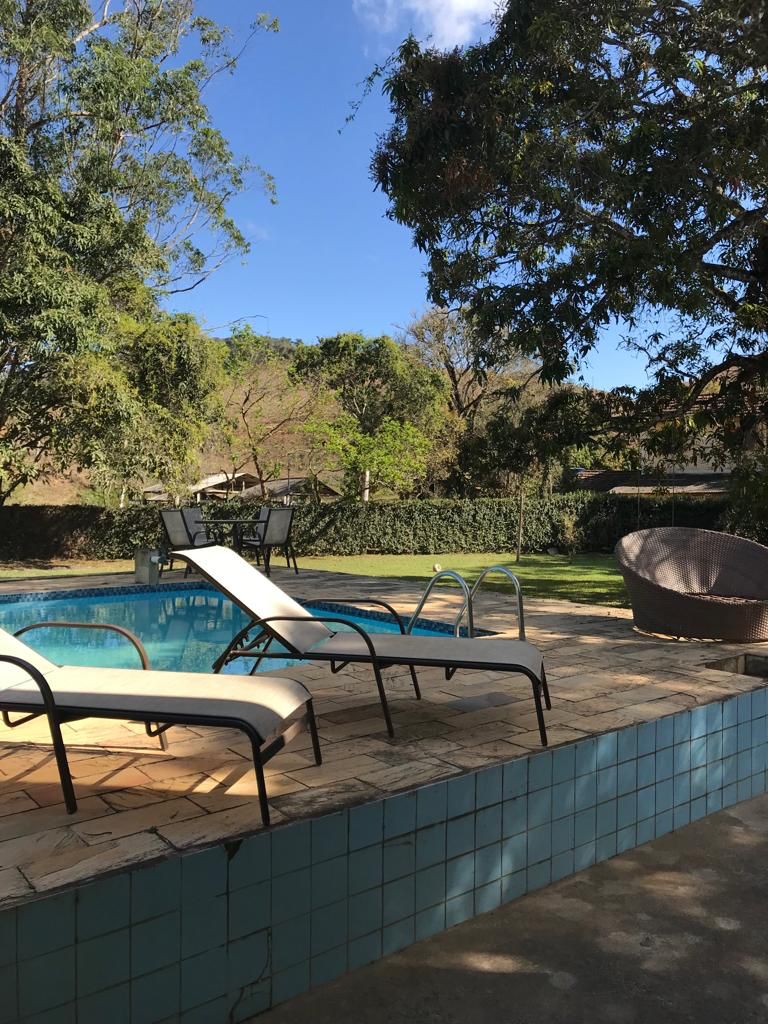 A swimming pool area is nestled among lush greenery, featuring lounge chairs positioned on a tiled deck. Light filtering through trees creates a serene atmosphere. A shaded dining area with tables is visible in the distance, complementing the outdoor relaxation space.