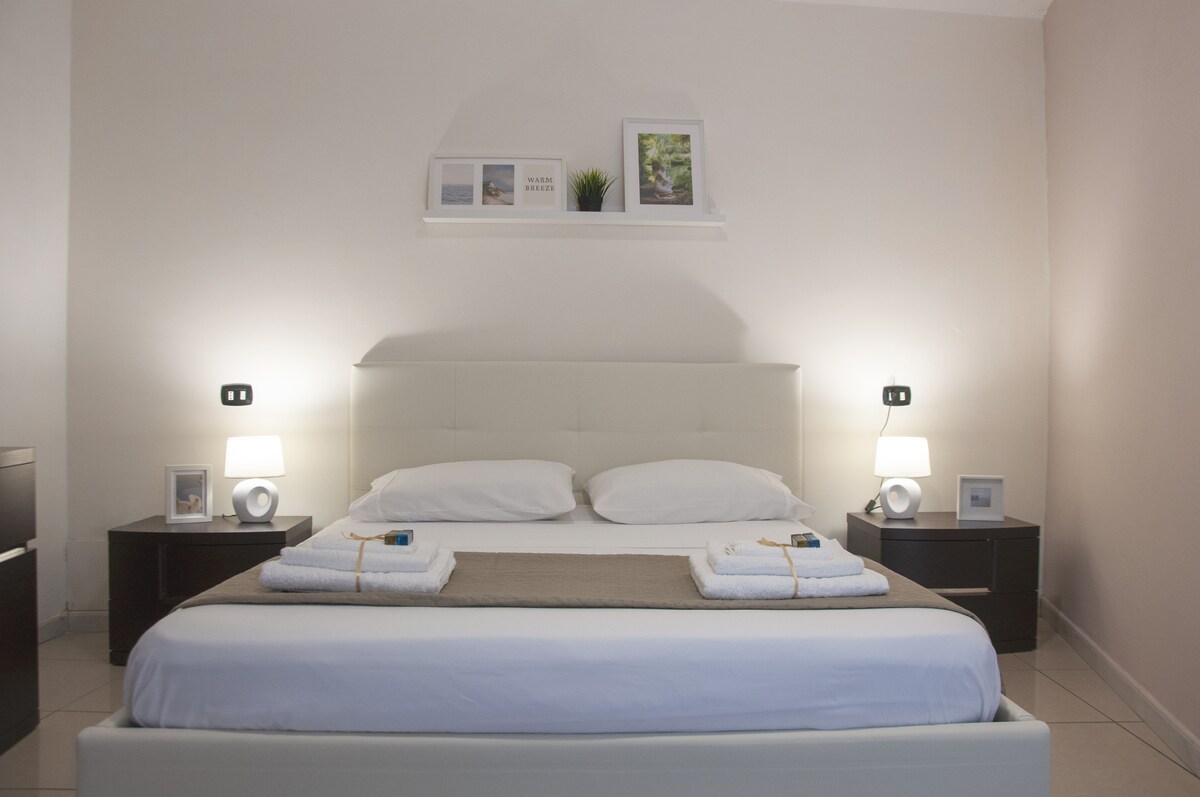 A spacious bedroom features a light-colored bed with neatly arranged linens and towels. Nightstands are placed on either side of the bed, each holding a lamp and framed photos. Soft lighting creates a calm environment, complemented by neutral wall tones.