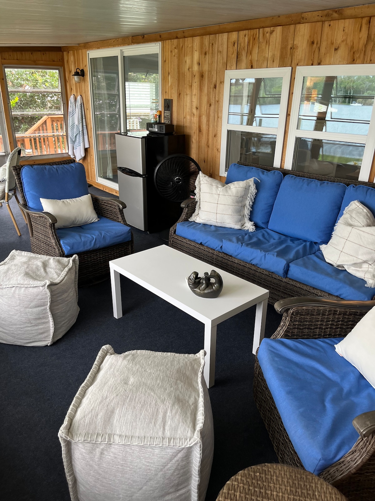 A cozy sunroom features a seating arrangement with wicker chairs and a matching loveseat, adorned with blue cushions. A white coffee table sits at the center, complemented by two light-colored poufs. Large windows provide views of the outdoor area, enhancing the inviting atmosphere.