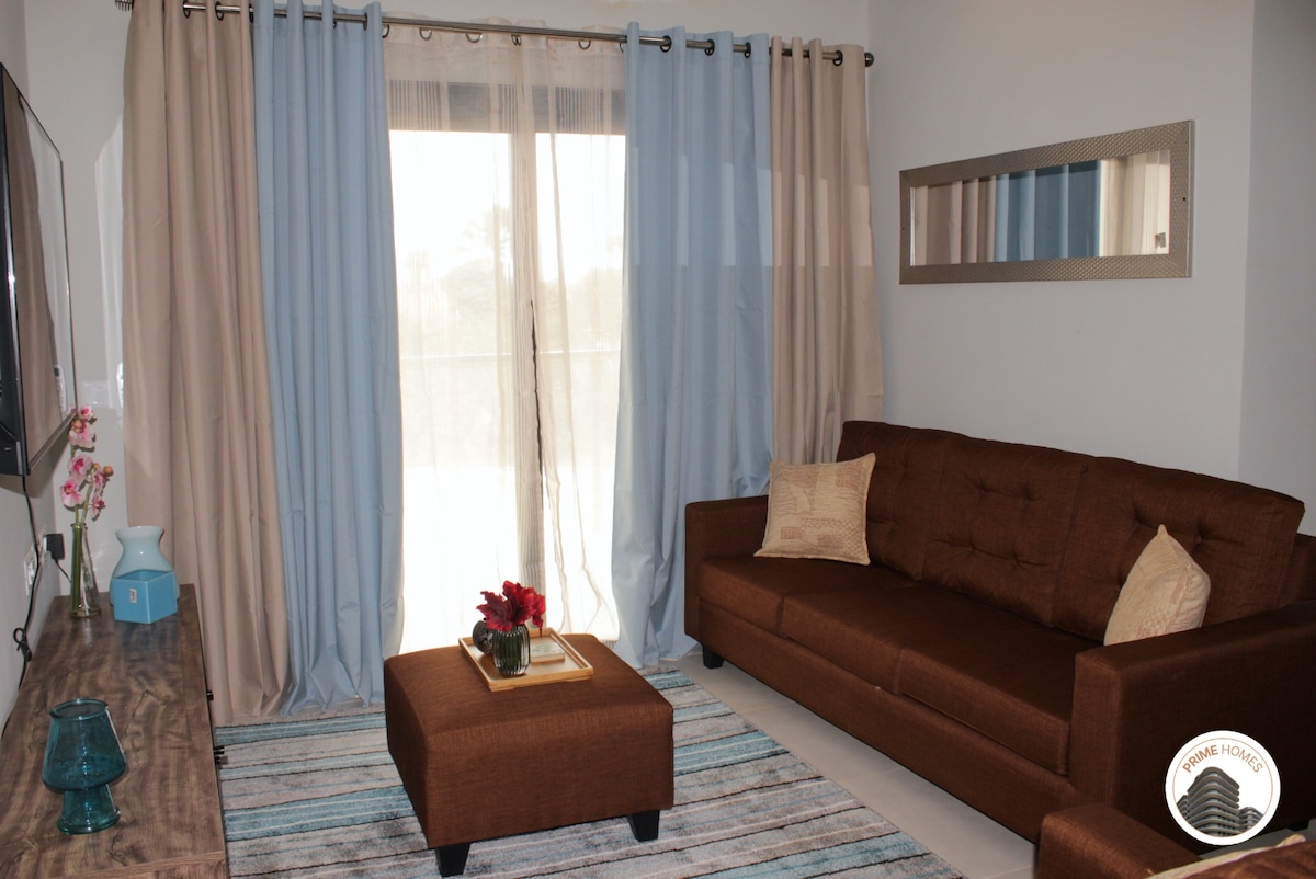 A comfortable sitting area features a brown upholstered sofa and an ottoman, complemented by a wooden coffee table. Soft blue and beige curtains frame the sliding glass doors that lead to the balcony, allowing natural light to fill the space.