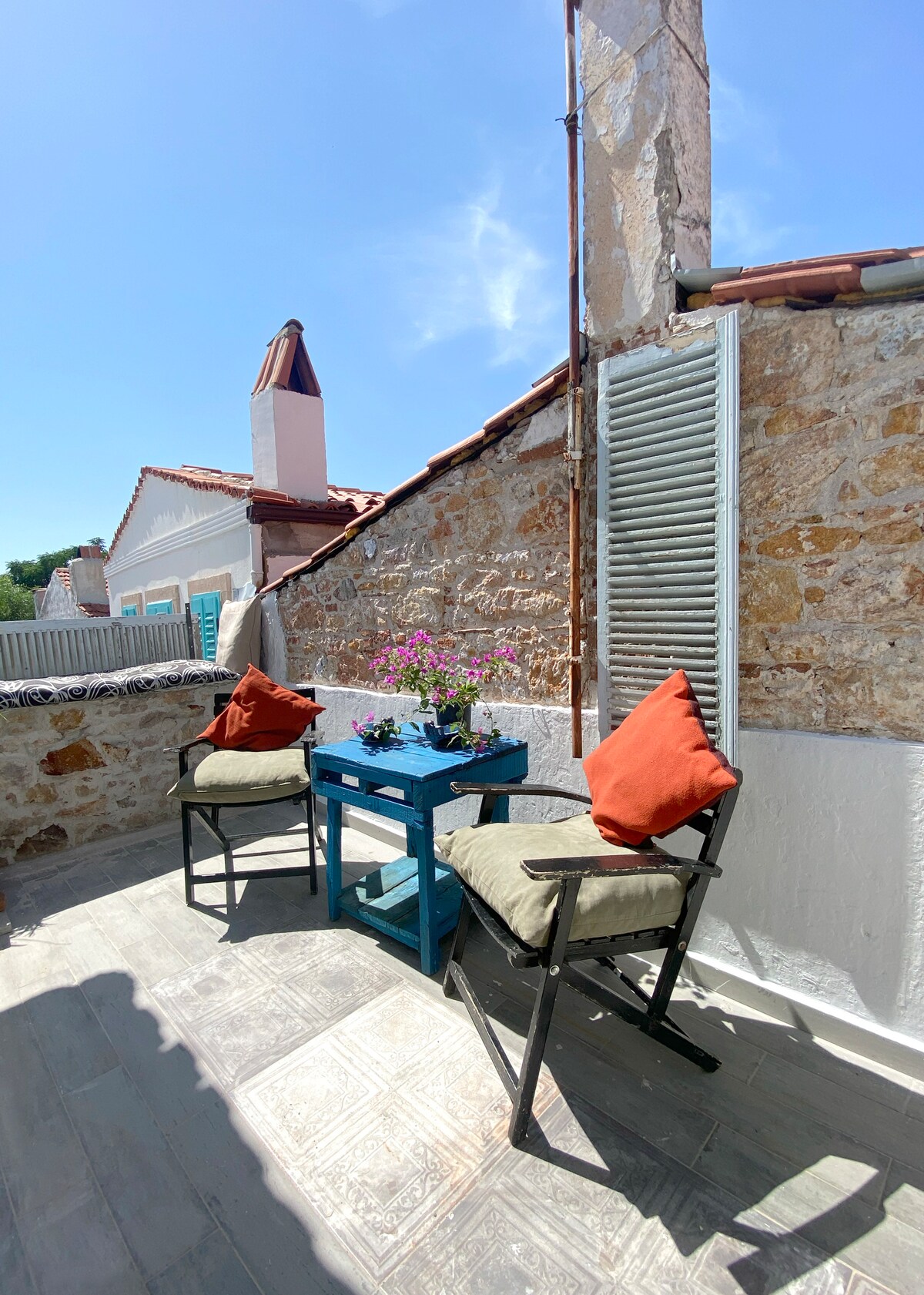 A small balcony features two tall chairs with cushions and a blue table holding a flower pot filled with vibrant blooms. The stone walls and charming shutters contribute to the cozy outdoor space, ideal for relaxation.