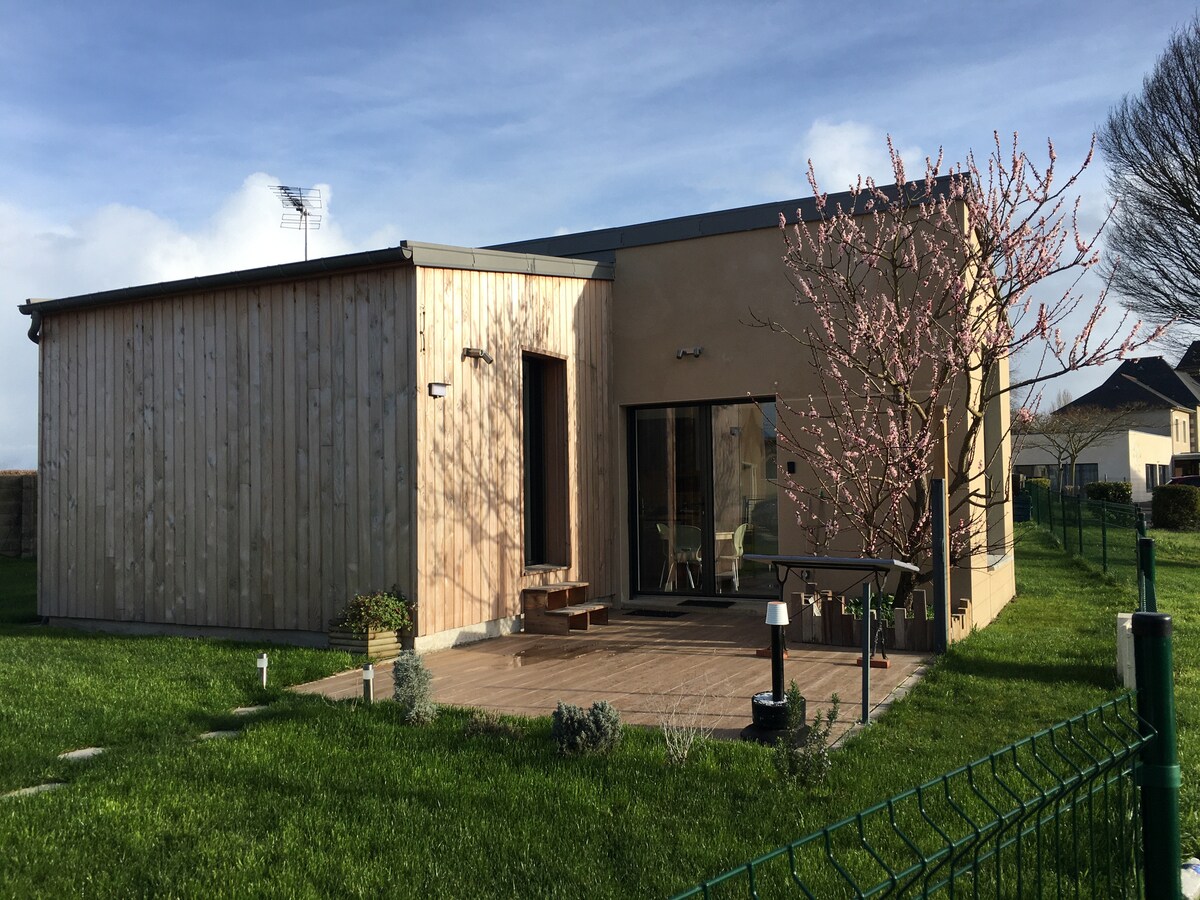 A modern exterior of a petite maison is presented, featuring wooden panels and a stone finish. A patio area is visible, surrounded by a well-maintained green lawn. Flowering trees and shrubs add greenery, while a secure fence encloses the property, enhancing privacy.