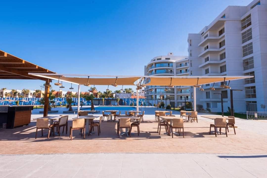 An outdoor dining area features several wood-framed tables and chairs positioned under a large shaded canopy. The blue water of the nearby pool reflects the clear sky, with resort buildings visible in the background, showcasing a relaxed atmosphere ideal for leisure.