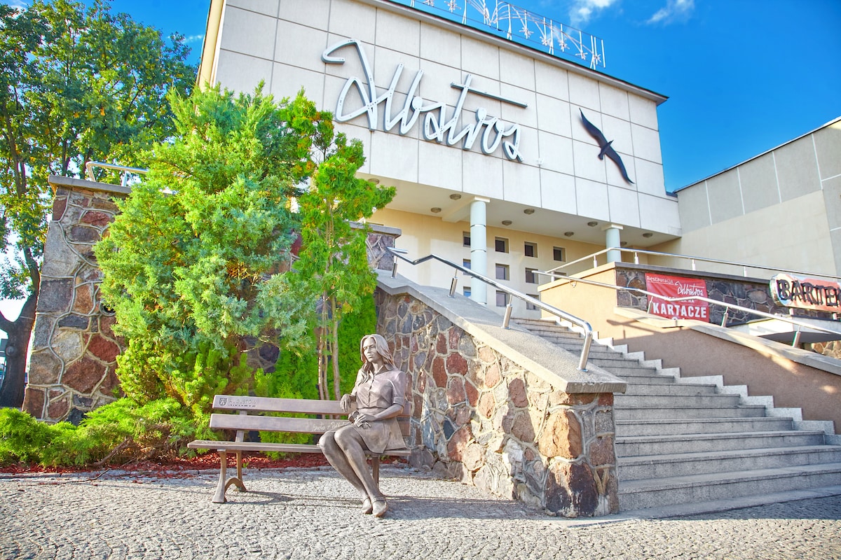 The exterior of the Albatros building features a prominent sign and stone steps leading to the entrance. A landscaped area with trees and a statue on a bench adds to the welcoming ambiance. Clear blue skies are visible above, enhancing the inviting setting.