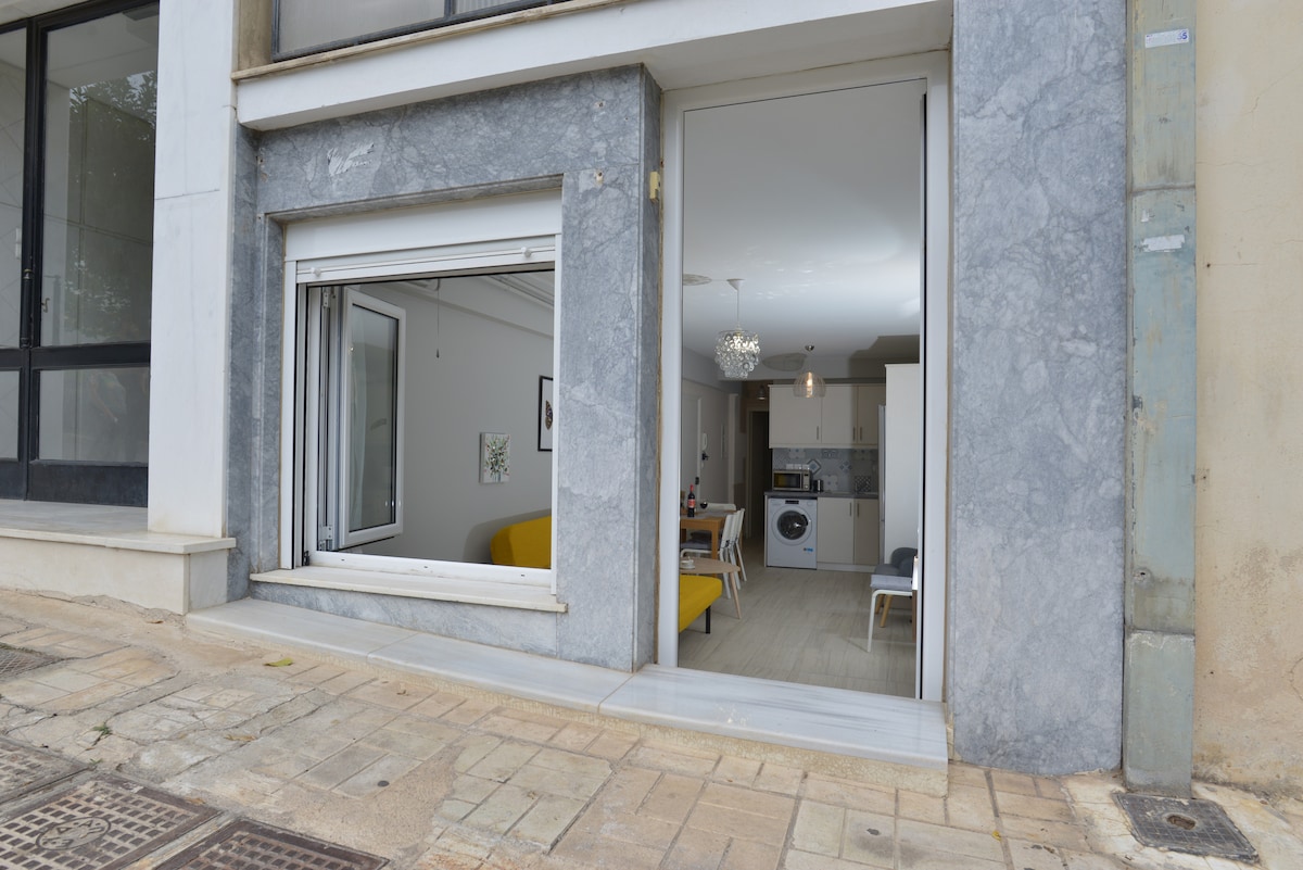 The exterior view of the apartment reveals a separate entrance framed by sleek grey stone. A large window opens to a bright interior, showcasing the living space, with visible kitchen components and a glimpse of a cozy sofa. The pedestrian street environment adds to the accessibility.