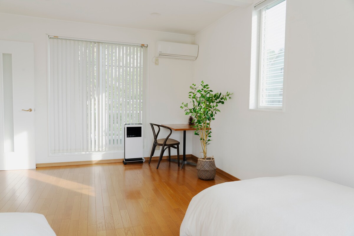 A bright and airy sleeping area is presented with light wooden flooring. A spacious bed is visible, accompanied by a simple desk and chair near a window. Soft natural light filters through vertical blinds, and a potted plant adds a touch of greenery.