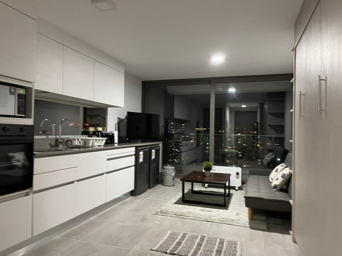 A modern kitchen area is equipped with stainless steel appliances, including an oven and a refrigerator. A sleek dining table is positioned beside a cozy seating area with a sofa. Large windows offer city views while light fixtures provide ample illumination.