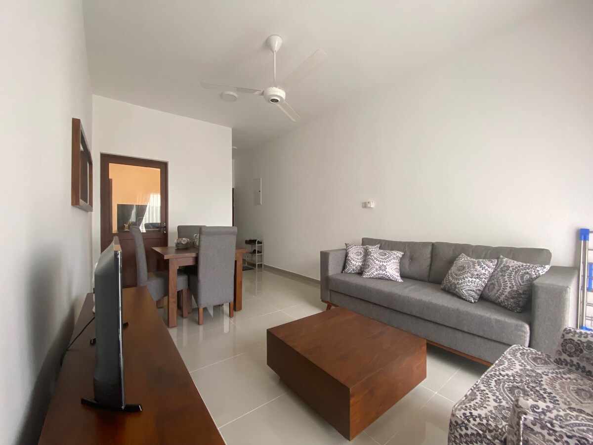 A modern living area features a gray sofa and a wooden coffee table. An armchair with a patterned design complements the space. A dining table with four chairs is positioned near a wall, and a flat-screen TV is visible across the room.