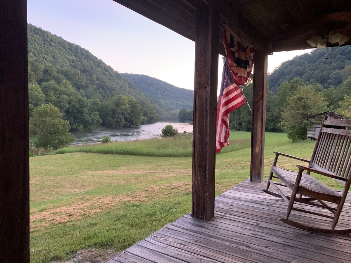 A view from the porch features a gently rolling landscape, with the Clinch River flowing through lush greenery. A wooden rocking chair invites relaxation, while an American flag hangs nearby, fluttering softly in the breeze. The image captures a tranquil and secluded natural setting.