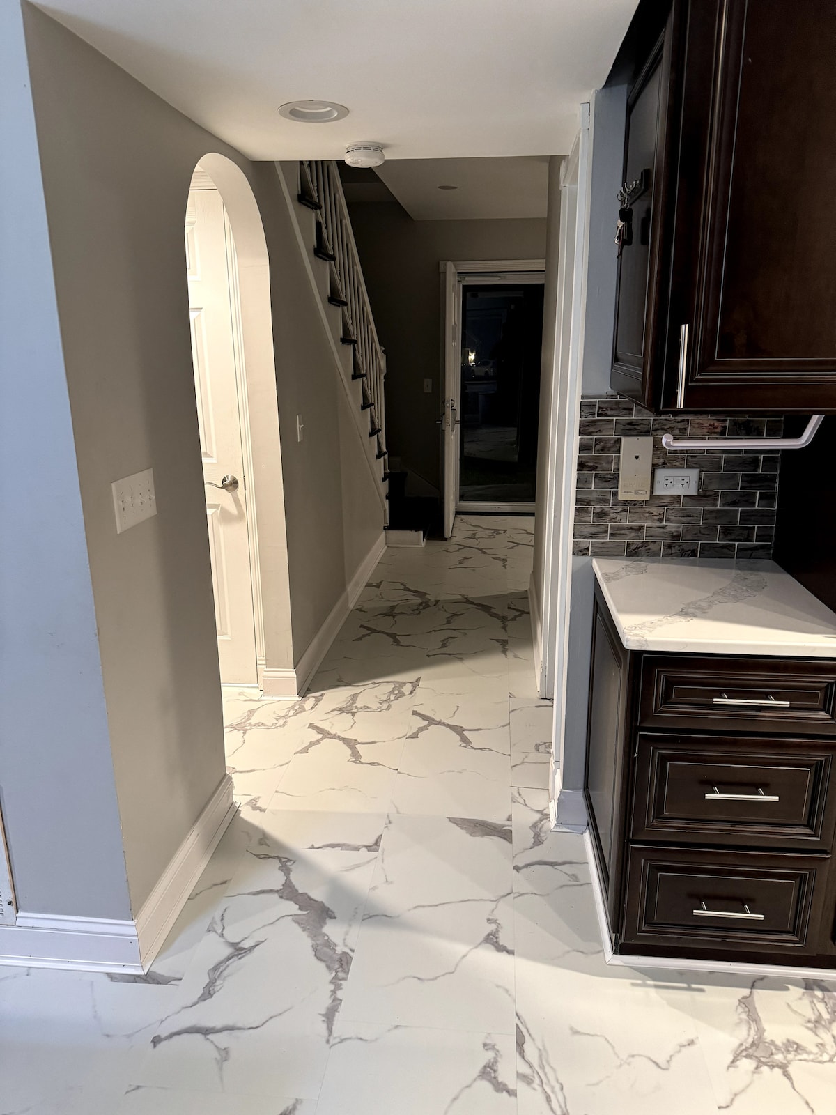 A hallway is featured, showcasing elegant marble-patterned flooring that leads toward a staircase. Light-colored walls contrast with dark cabinetry along one side. A doorway at the end opens to the outdoors, allowing natural light to illuminate the space.
