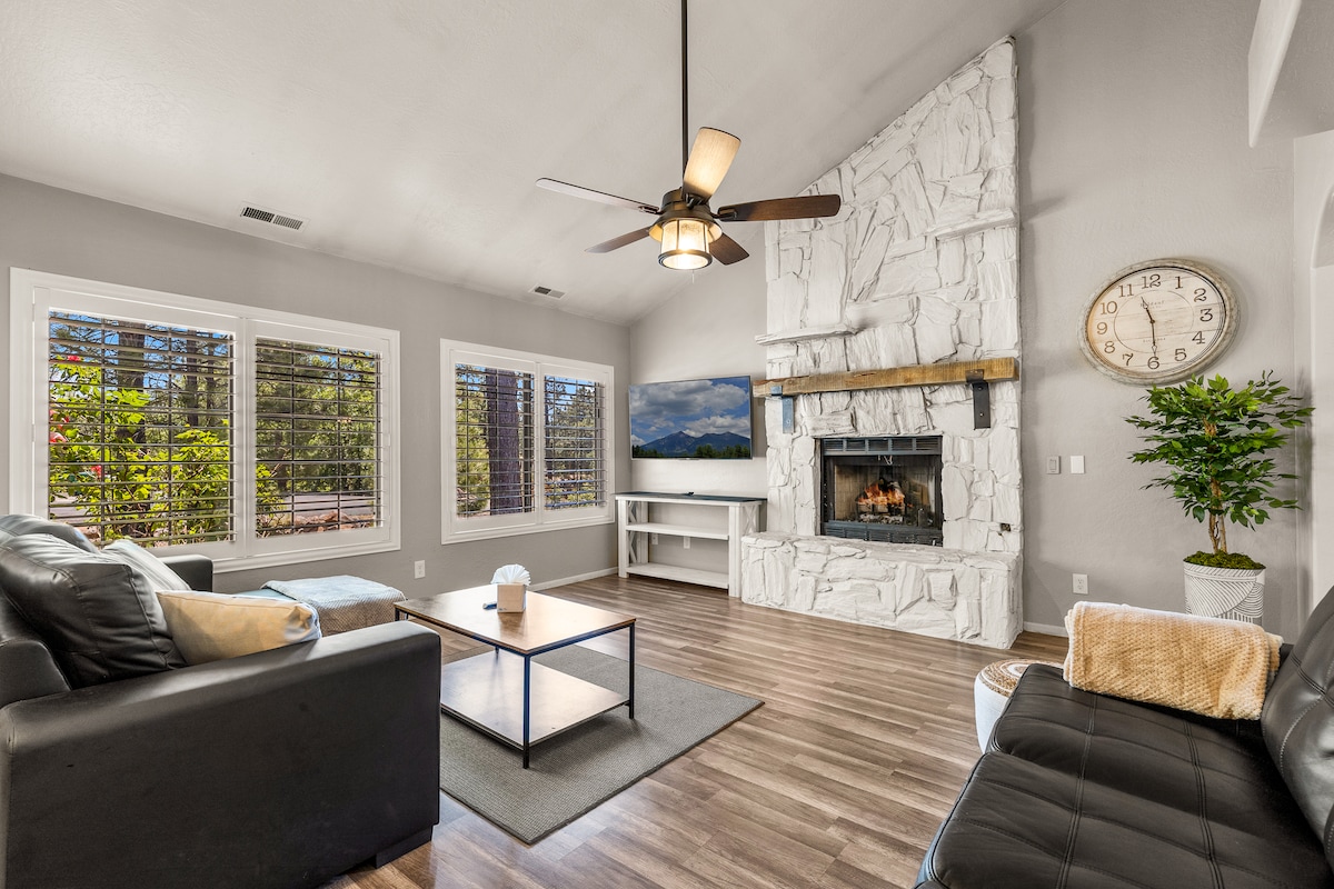 A spacious living room features comfortable seating arranged around a central coffee table. A stone fireplace contrasts with the light walls, while large windows invite natural light and views of surrounding greenery. A television is mounted on the wall opposite the seating area.