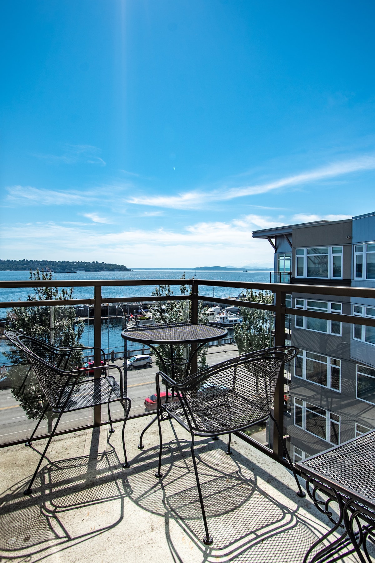 A private balcony is showcased with a small round table and two matching chairs, offering a direct view of Elliott Bay. The water sparkles under bright blue skies, with the marina and distant shoreline visible. Natural light enhances the space, surrounded by greenery.
