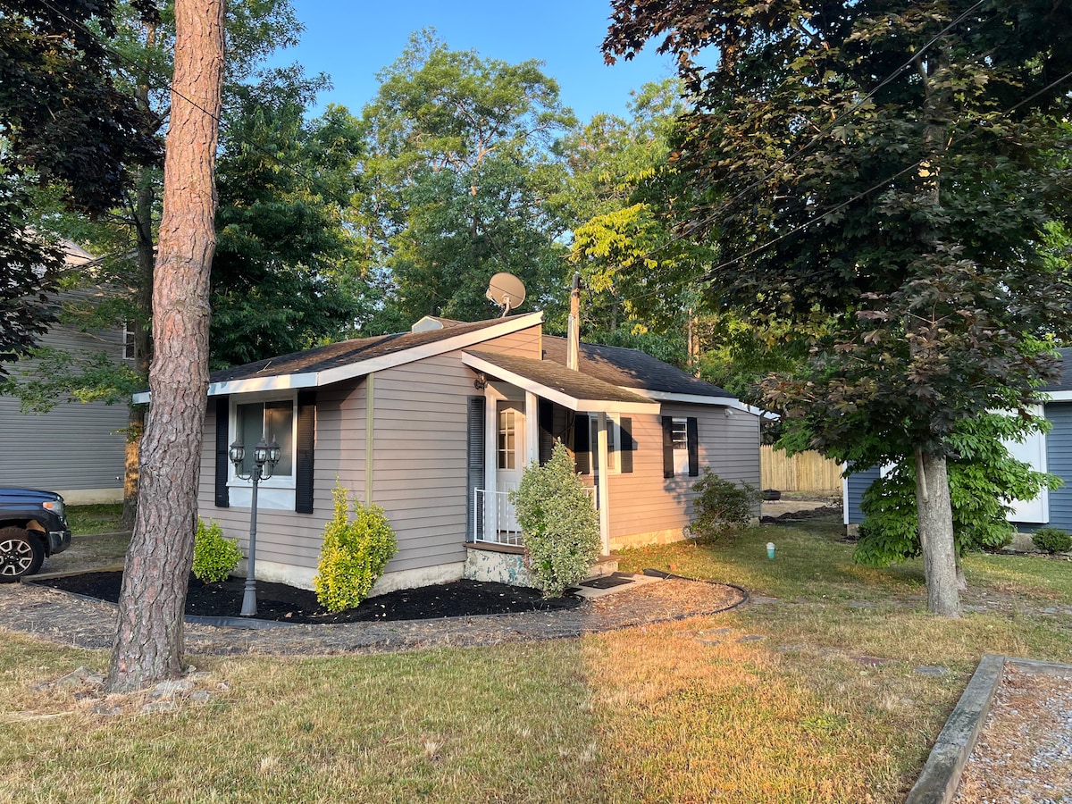 A single-story house is surrounded by lush greenery, with tall trees providing shade. The exterior features light-colored siding complemented by black shutters. A well-maintained lawn and gravel pathway lead to the entrance, creating a welcoming appeal in a peaceful neighborhood.
