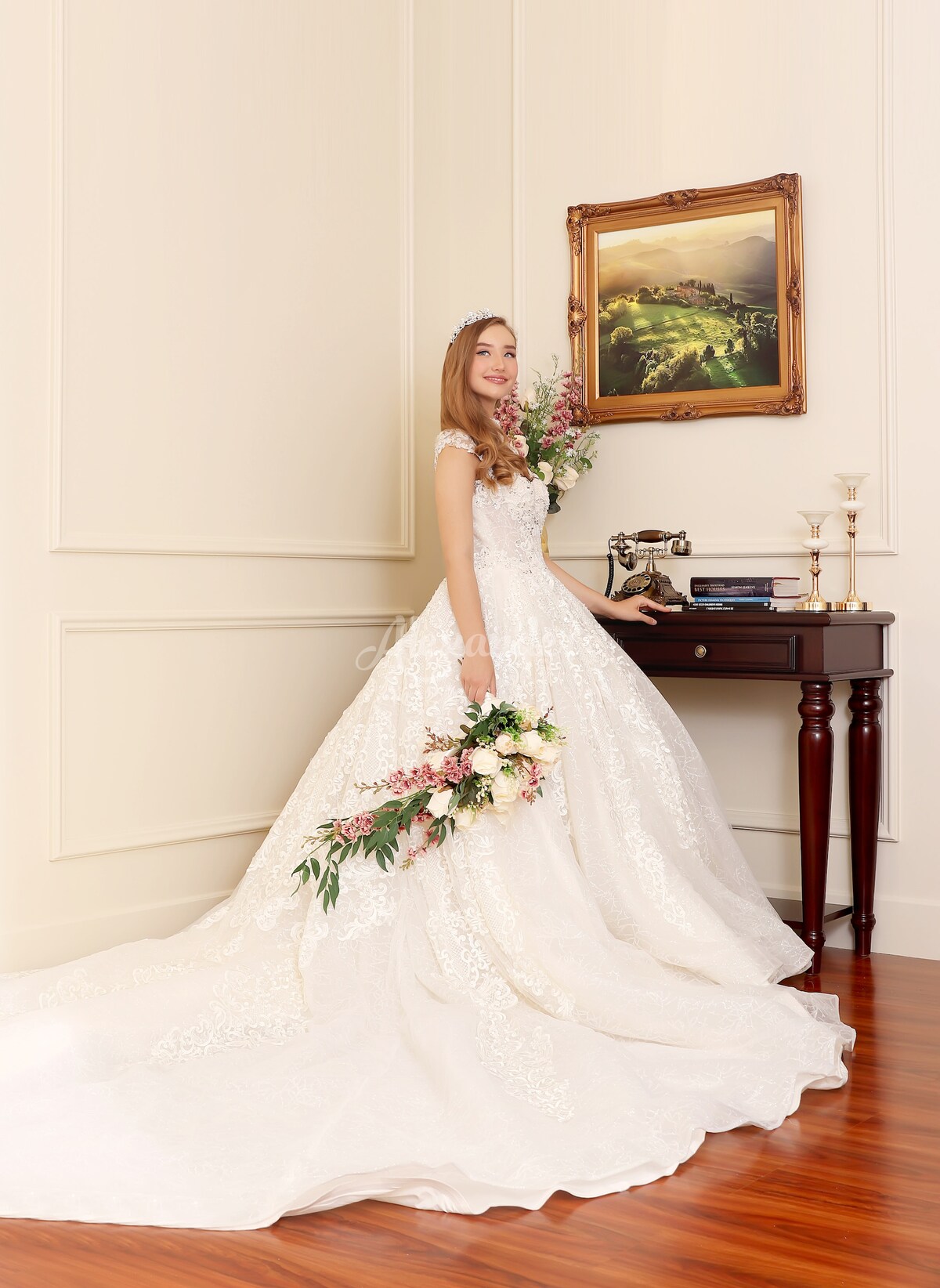 A wedding gown adorned with intricate lace details is showcased in an elegant setting. The dress flows gracefully to the floor, while a bouquet of flowers complements its design. A vintage sewing machine and an ornate framed painting are positioned on a wooden table nearby.