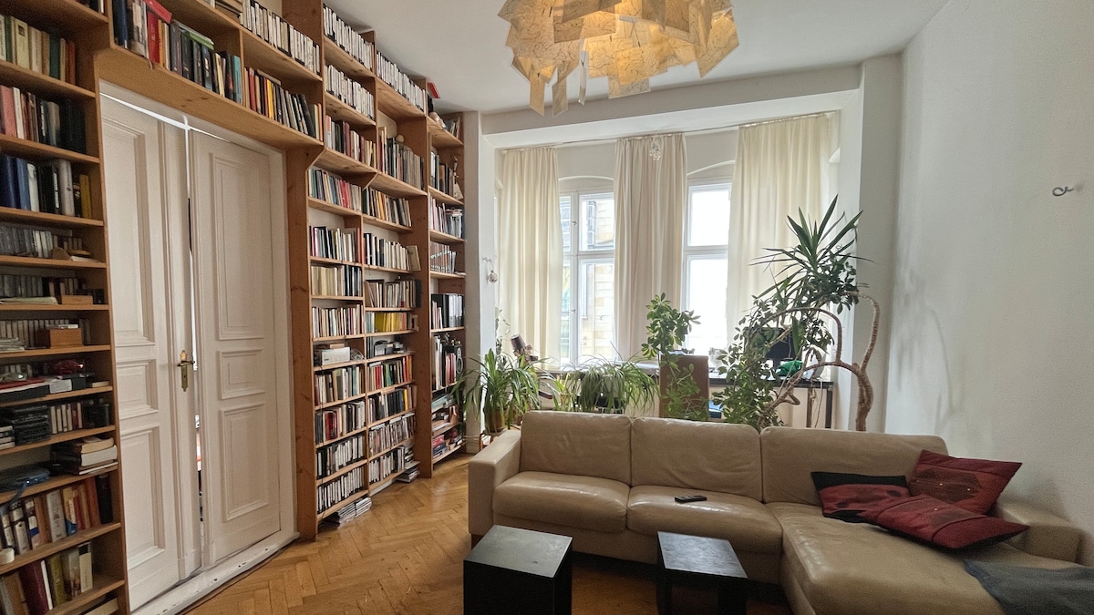 A cozy corner is filled with a large bookshelf showcasing a variety of books. A light-colored couch sits opposite a coffee table, surrounded by greenery from potted plants. Natural light filters through large windows, softly illuminating the space.