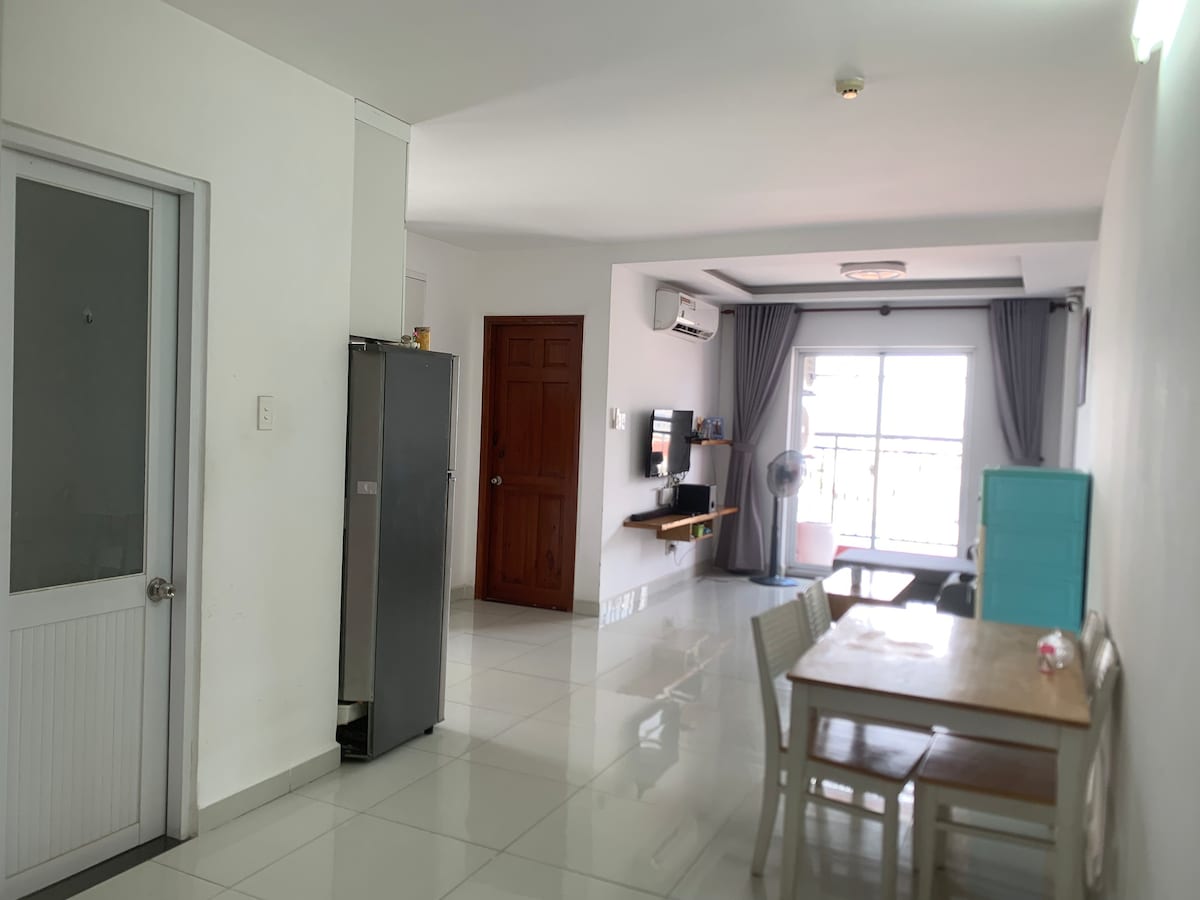 A bright open area is shown, featuring a dining table with chairs and a kitchen area with a refrigerator. A living space is visible, equipped with a television and a fan, with sliding glass doors leading to a balcony. Natural light fills the room.