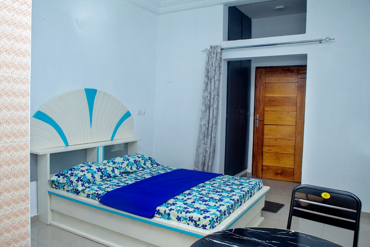 A well-appointed sleeping area is highlighted by a floral-patterned bedspread on a spacious bed. A blue blanket is neatly placed over the bed. The room features light blue walls, a closed wooden door, and a small dining table with a black chair.