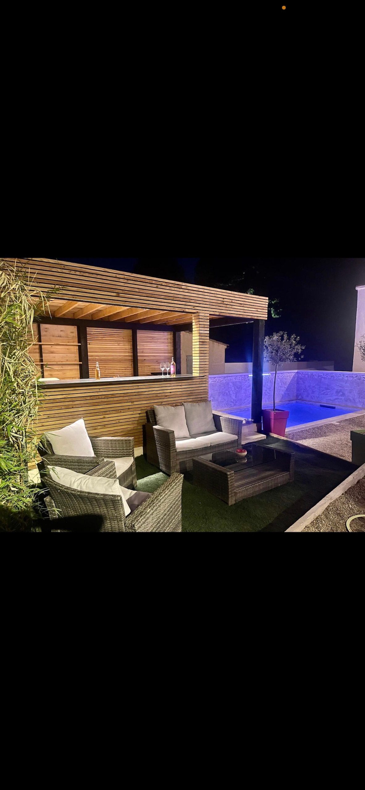 An outdoor seating area features comfortable wicker chairs and a table, surrounded by wooden slats for privacy. The illuminated pool in the background reflects soft light, enhancing the ambiance. Planters with greenery add a natural touch to the relaxing space.