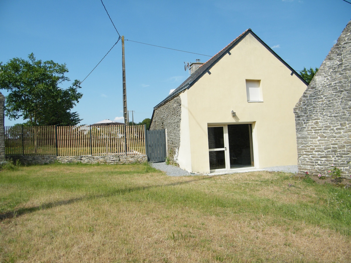A single-story stone cottage is presented alongside a grassy yard. The front features large glass doors that open to the outdoor space, while a wooden fence surrounds the property. A clear blue sky and sparse greenery enhance the peaceful environment.