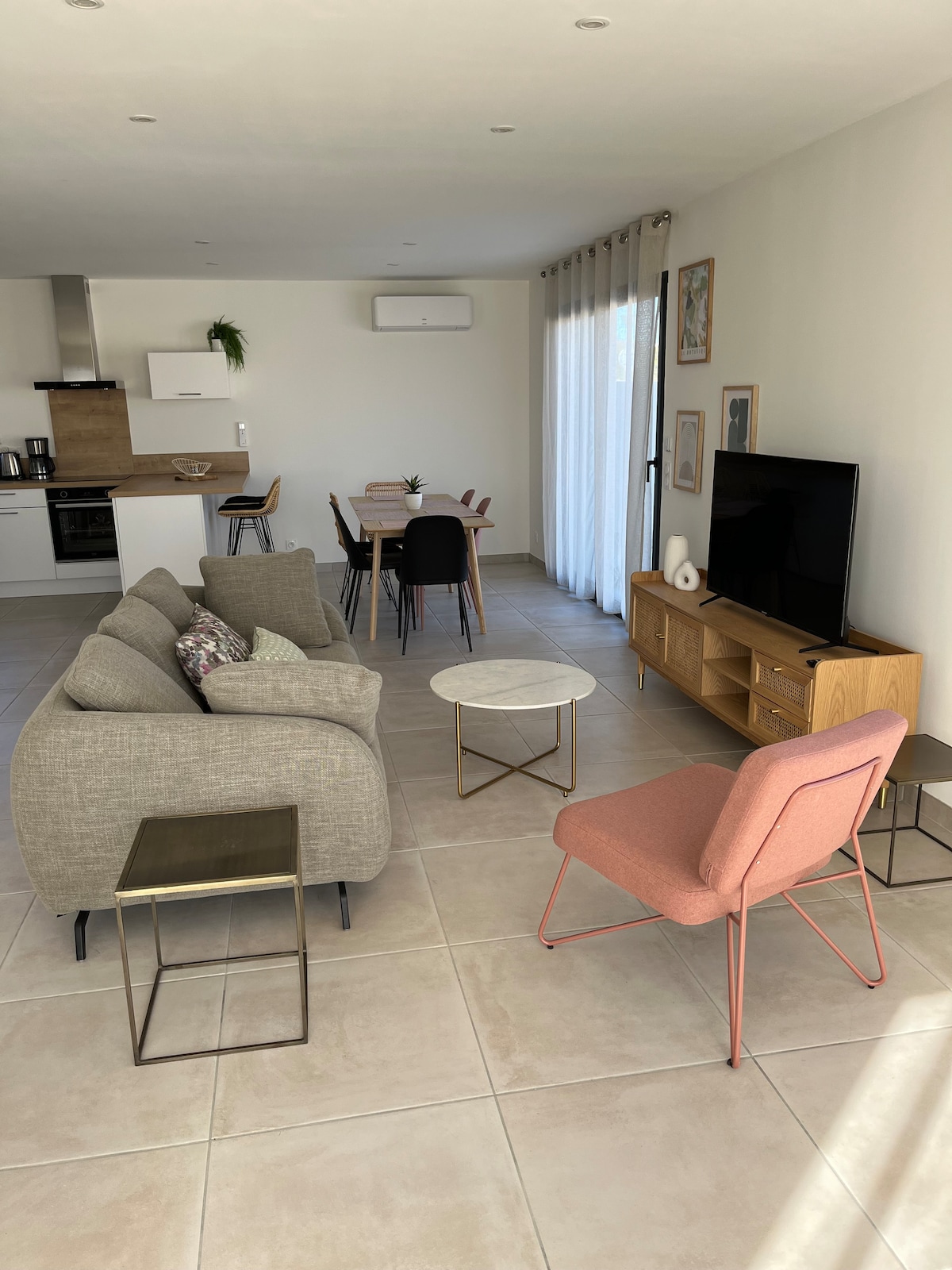 The open-plan living area features a light-filled space with a comfortable gray sofa, a pink accent chair, and a modern coffee table. A dining table for six sits nearby, and a flat-screen TV is positioned against the wall, providing a welcoming atmosphere for relaxation and dining.