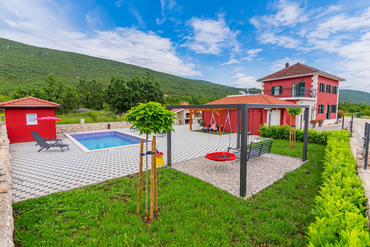The image shows a vibrant red villa surrounded by greenery, featuring a private pool and a children's playground with swings. A spacious patio with lounge chairs complements the outdoor space, while the backdrop showcases rolling hills under a clear blue sky.