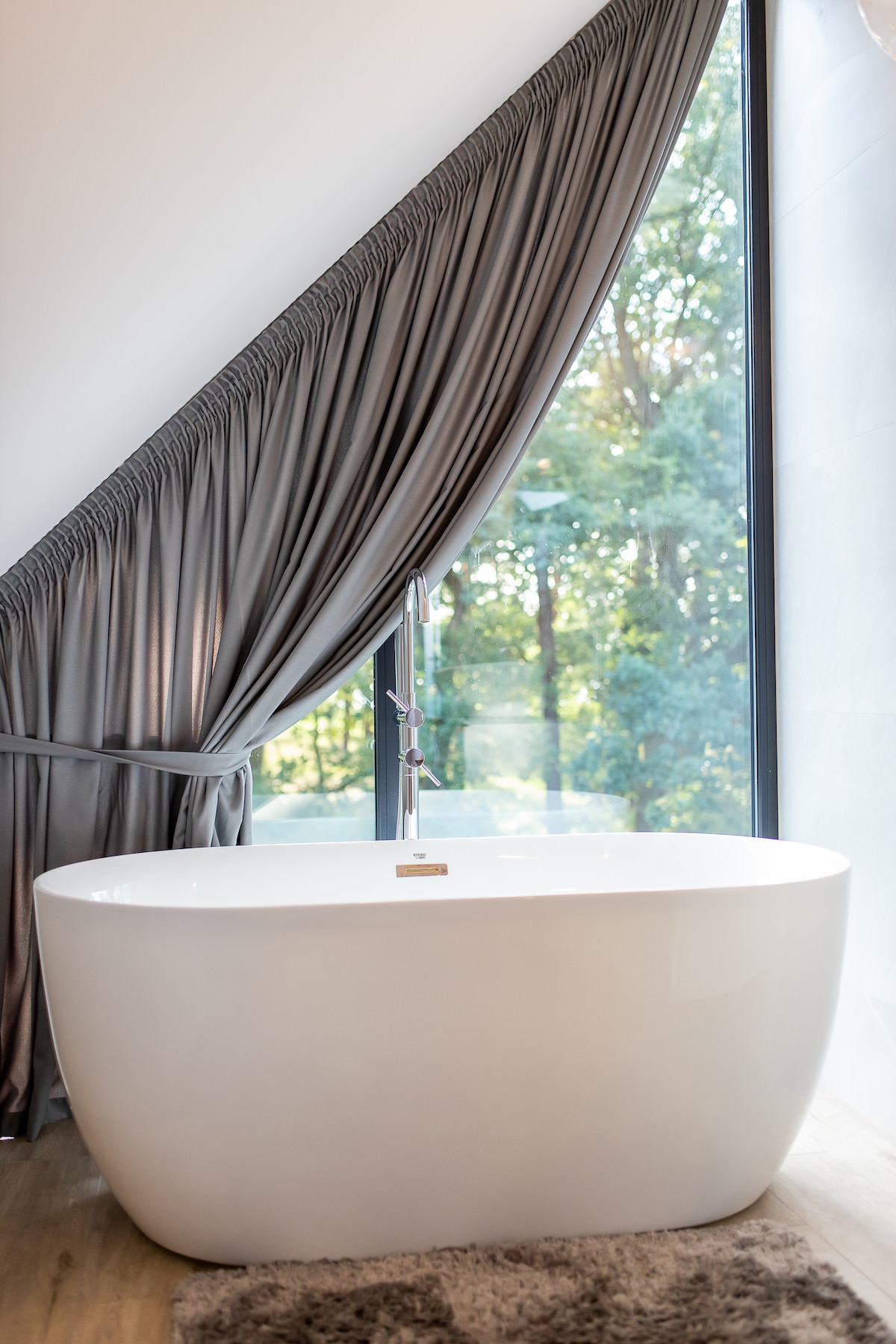 A freestanding bathtub is positioned beneath a large window, allowing natural light to illuminate the space. Soft drapery frames the window, while greenery can be seen outside, enhancing a serene ambiance. A plush rug lies on the floor, providing comfort underfoot.