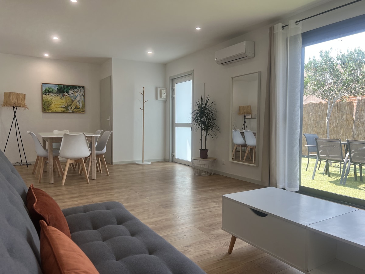 A bright living area features a comfortable gray sofa adorned with orange cushions. Natural light enters through large windows, illuminating the space. A dining table with white chairs and a potted plant adds a welcoming touch, while a mirror reflects the room’s ambiance.
