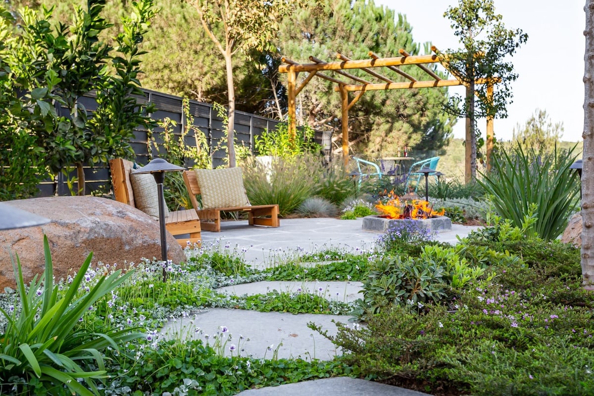 A peaceful garden area is presented with stone pathways leading to a fire pit, surrounded by varied greenery and flowering plants. Comfortable seating options are positioned nearby under a wooden trellis, offering a serene outdoor space for relaxation.