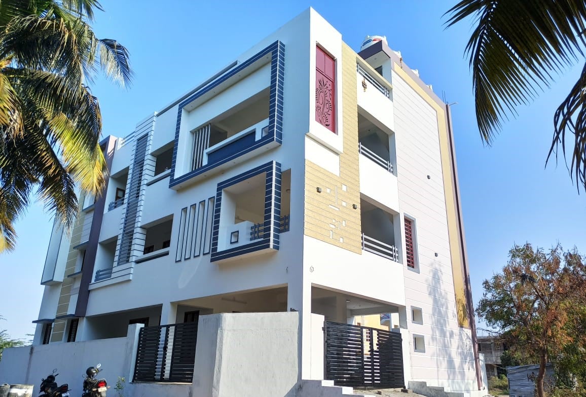 The exterior of the building is shown, featuring a modern design with a combination of light and dark tones. Large windows and multiple balconies create an open look. Lush palm trees are visible beside the entrance, enhancing the tranquil environment of the homestay.