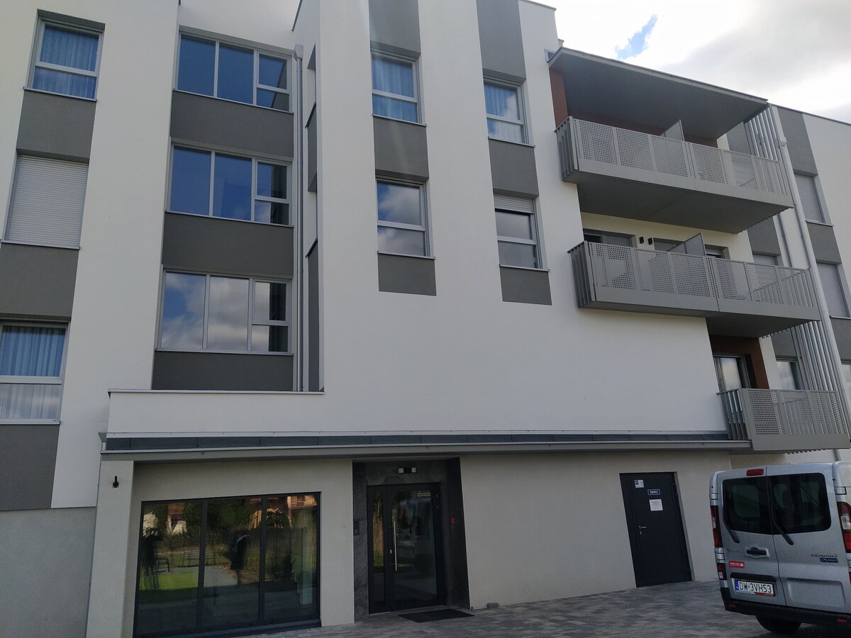 The contemporary building features a sleek facade with large windows allowing natural light to fill the interior. Balconies provide private outdoor space, while the main entrance is highlighted by glass doors and a paved area that enhances accessibility.