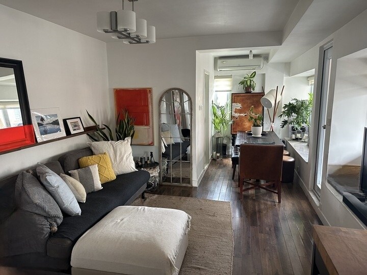 A bright living area is shown with a comfortable dark sofa adorned with decorative pillows. A stylish coffee table and a dining area with a chair complement the setting. Natural light enters through large windows, enhancing the plants and artwork displayed throughout the space.
