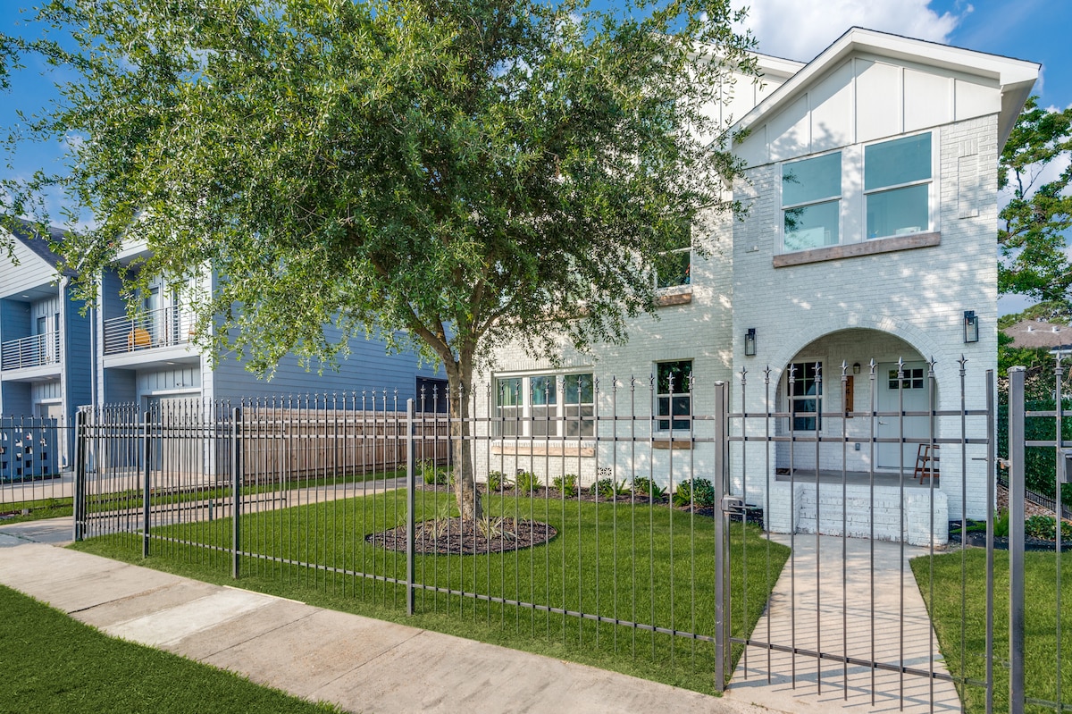 The exterior of a two-story building is framed by a low iron fence and a tree with lush green foliage. A pathway leads to an arched entrance, showcasing ample windows that invite light into the interior.
