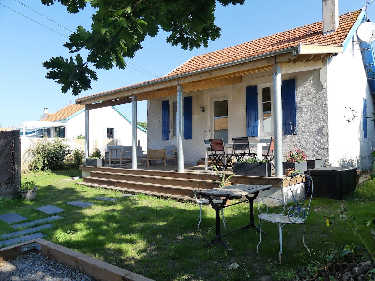 A charming exterior view of the house is presented, featuring a covered porch with wooden decking and blue shutters. A seating area and a dining table are visible on the porch, surrounded by green grass and garden elements. The welcoming entrance door is partially open, inviting guests inside.