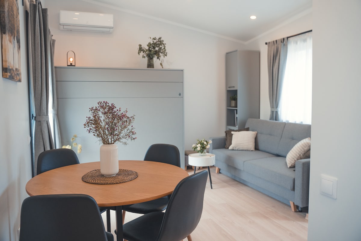 An open living area features a round wooden dining table surrounded by black chairs. A cozy sofa rests against the wall, complemented by a shelf with decorative elements. Natural light filters in through the curtains, creating a welcoming space.