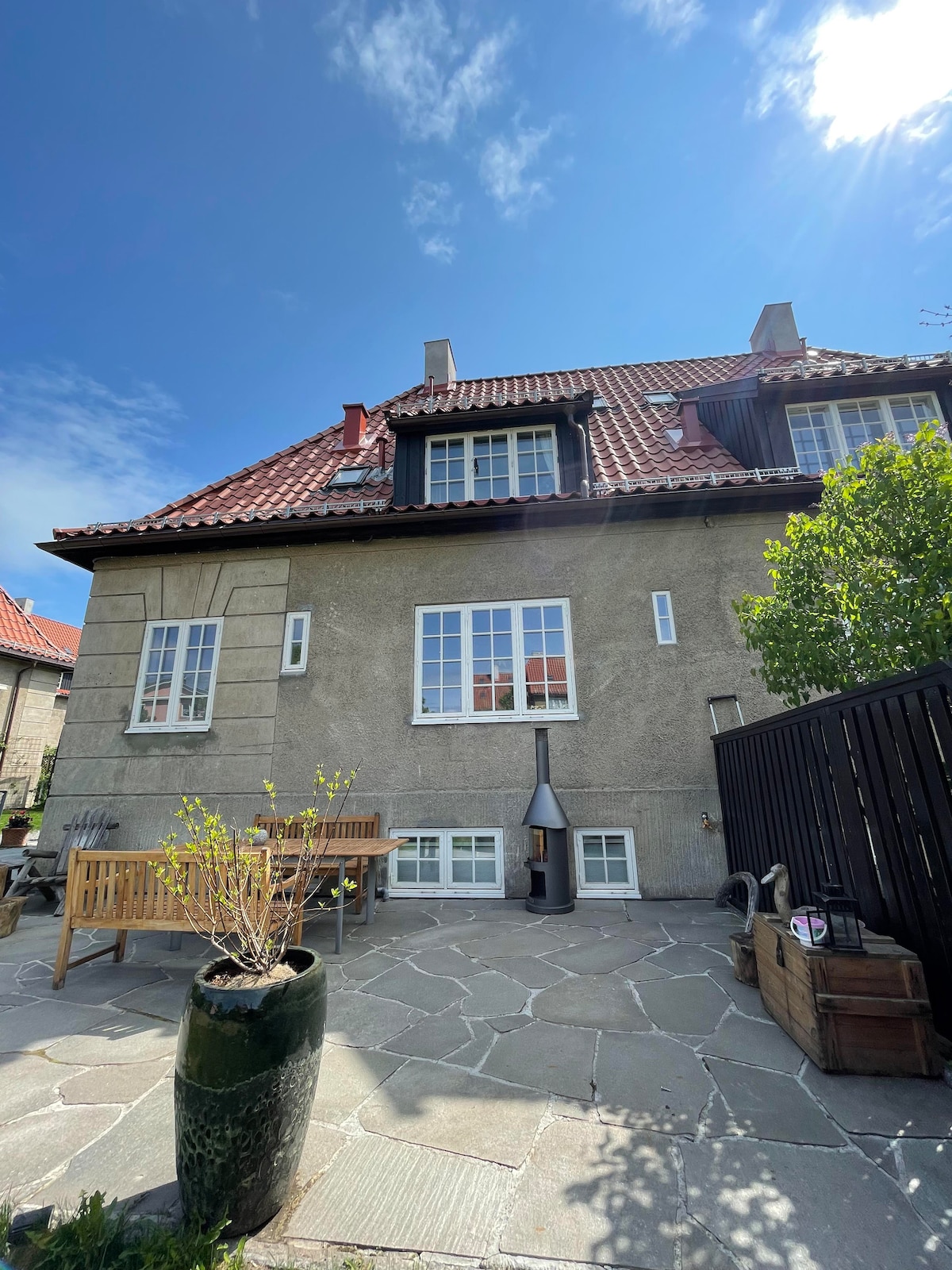 A charming exterior view of a two-story building is captured, featuring textured gray walls and red tile roofing. A small patio area with wooden benches is visible, accompanied by a stone pathway and greenery. Large windows allow natural light to brighten the facade.
